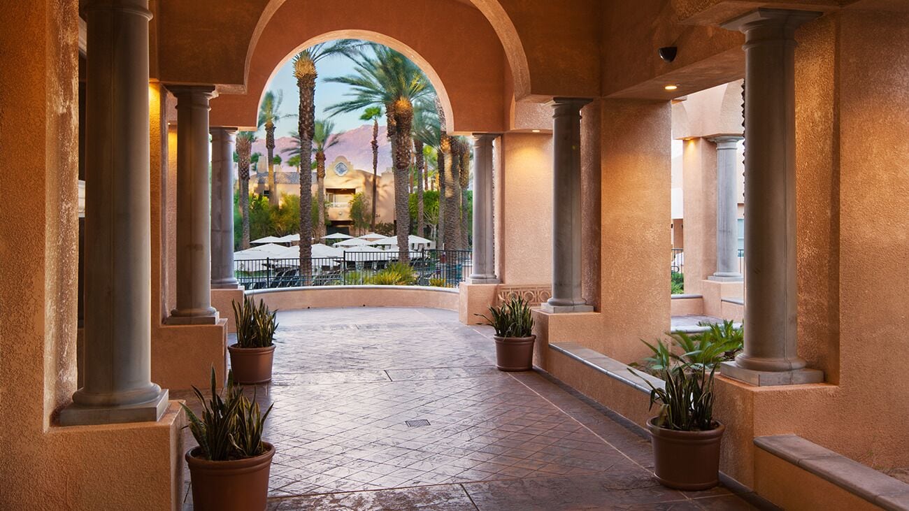 Un paseo techado con columnas, plantas en macetas y vistas a mesas al aire libre y palmeras.
