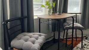 Un petit coin repas avec une table ronde et deux chaises, une plante en pot posée sur la table et une fenêtre avec des stores.