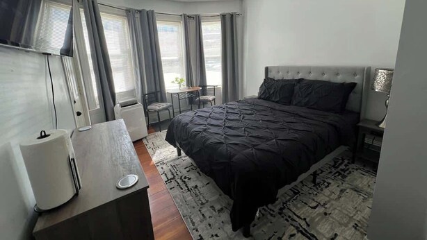Une chambre moderne avec un grand lit, un bureau et une chaise. On y trouve des fenêtres avec des rideaux, une petite table et une lampe.