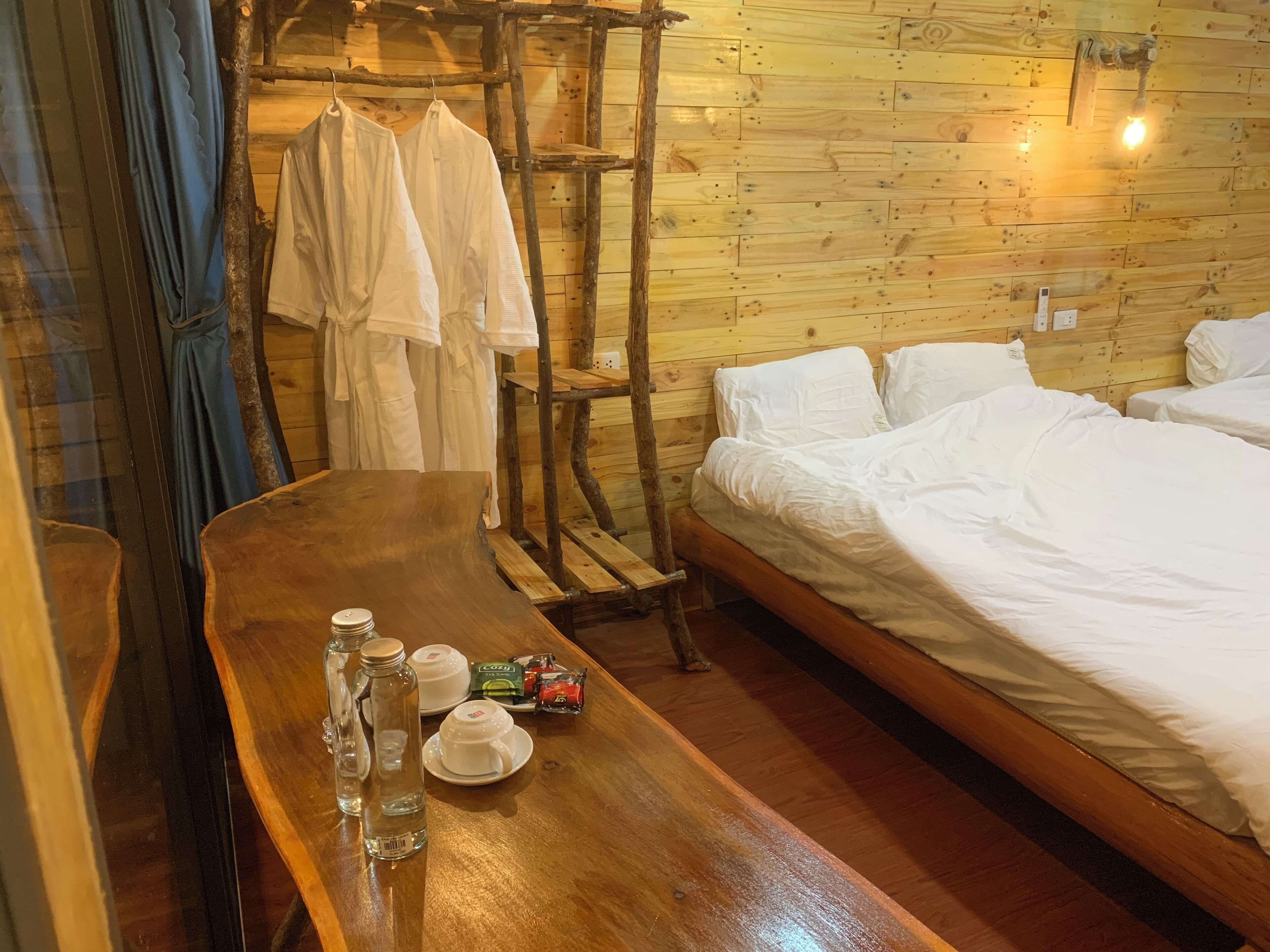 Ein hölzernes Bett mit weißen Leinen, ein Holztisch mit Flaschen und Tassen sowie ein Kleiderständer mit weißen Kleidungsstücken.