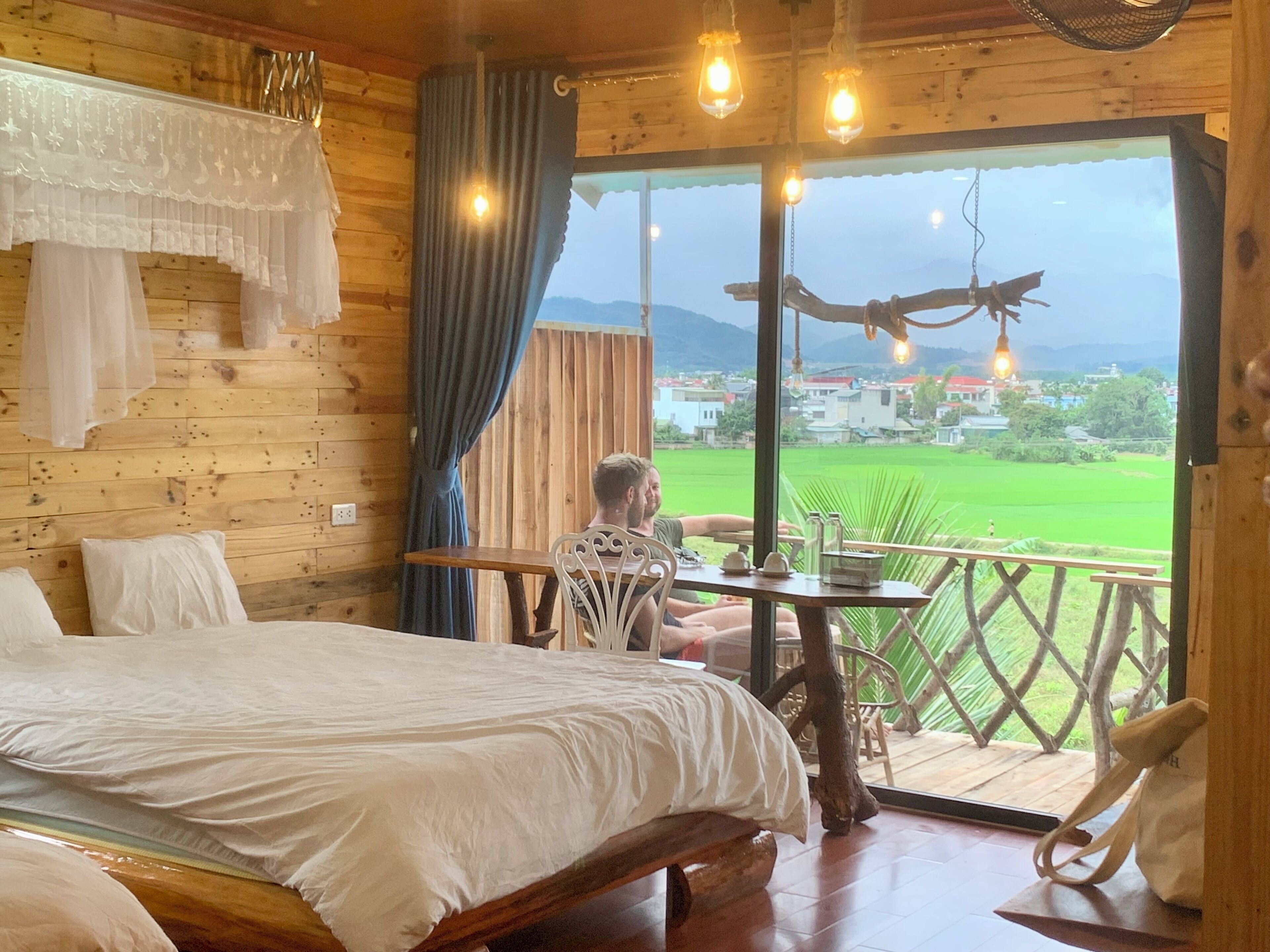 A wooden cabin room with a bed, a table, and a balcony with a view of a green field and mountains.