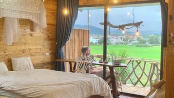 A wooden cabin room with a bed, a table, and a balcony with a view of a green field and mountains.