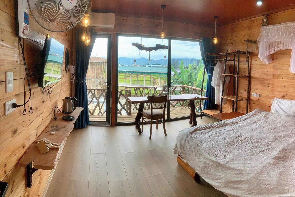 Een houten kamer met een bed, een tafel, een stoel, een ventilator en uitzicht op de natuur.