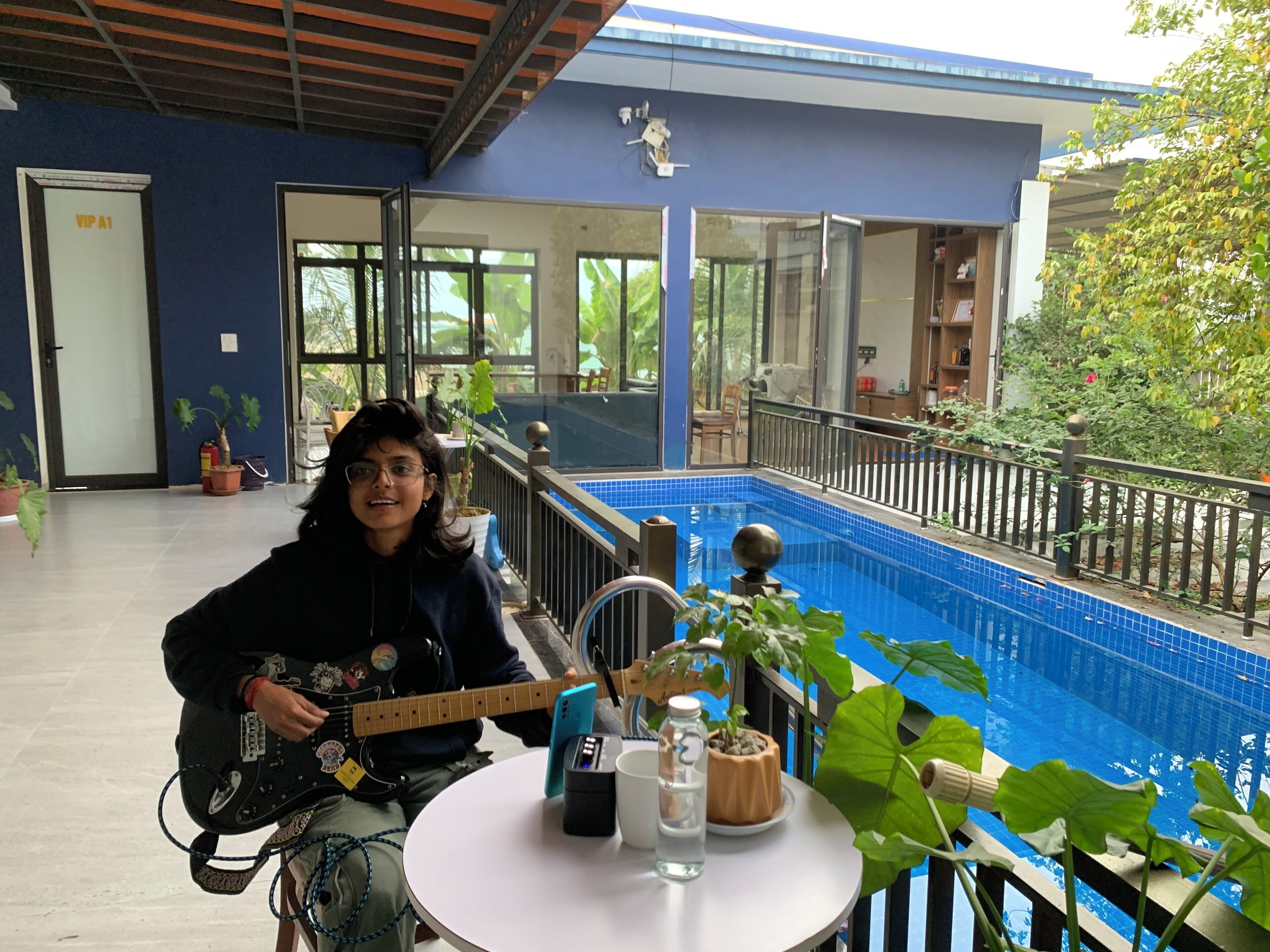 Una persona tocando la guitarra en una mesa con vista a una piscina, plantas en macetas y una piscina con azulejos azules.