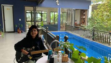 Una persona tocando la guitarra en una mesa con vista a una piscina, plantas en macetas y una piscina con azulejos azules.