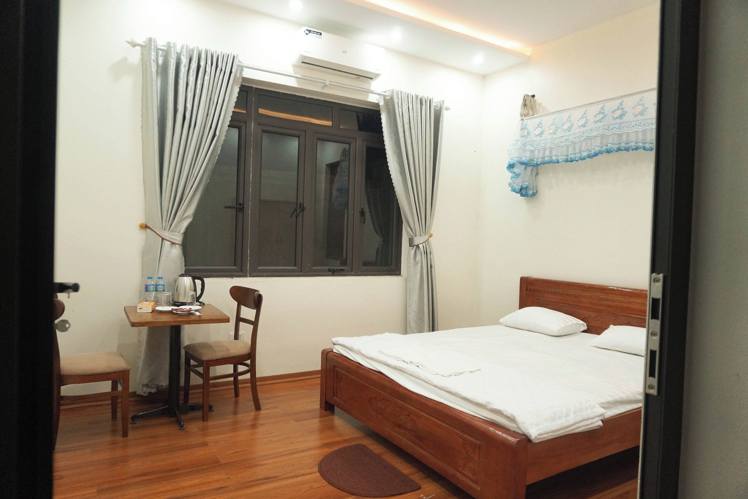 Um quarto com uma cama de madeira, uma escrivaninha com cadeira, uma janela com cortinas e uma prateleira fixada na parede.