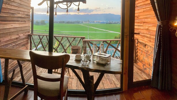 Un intérieur en bois avec une table et une chaise, une vue sur des champs verdoyants et des montagnes, et des luminaires suspendus.