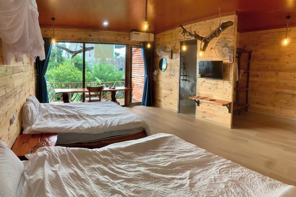 Een houten kamer met twee bedden, een tafel, stoelen, een douche en een balkon met uitzicht op groen.