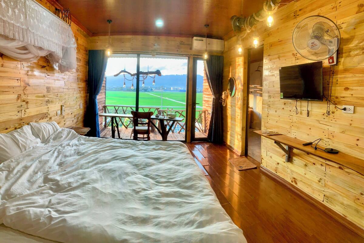 Een houten kamer met een bed, een tafel en een televisie. Er is een balkon met uitzicht op een veld en bergen.