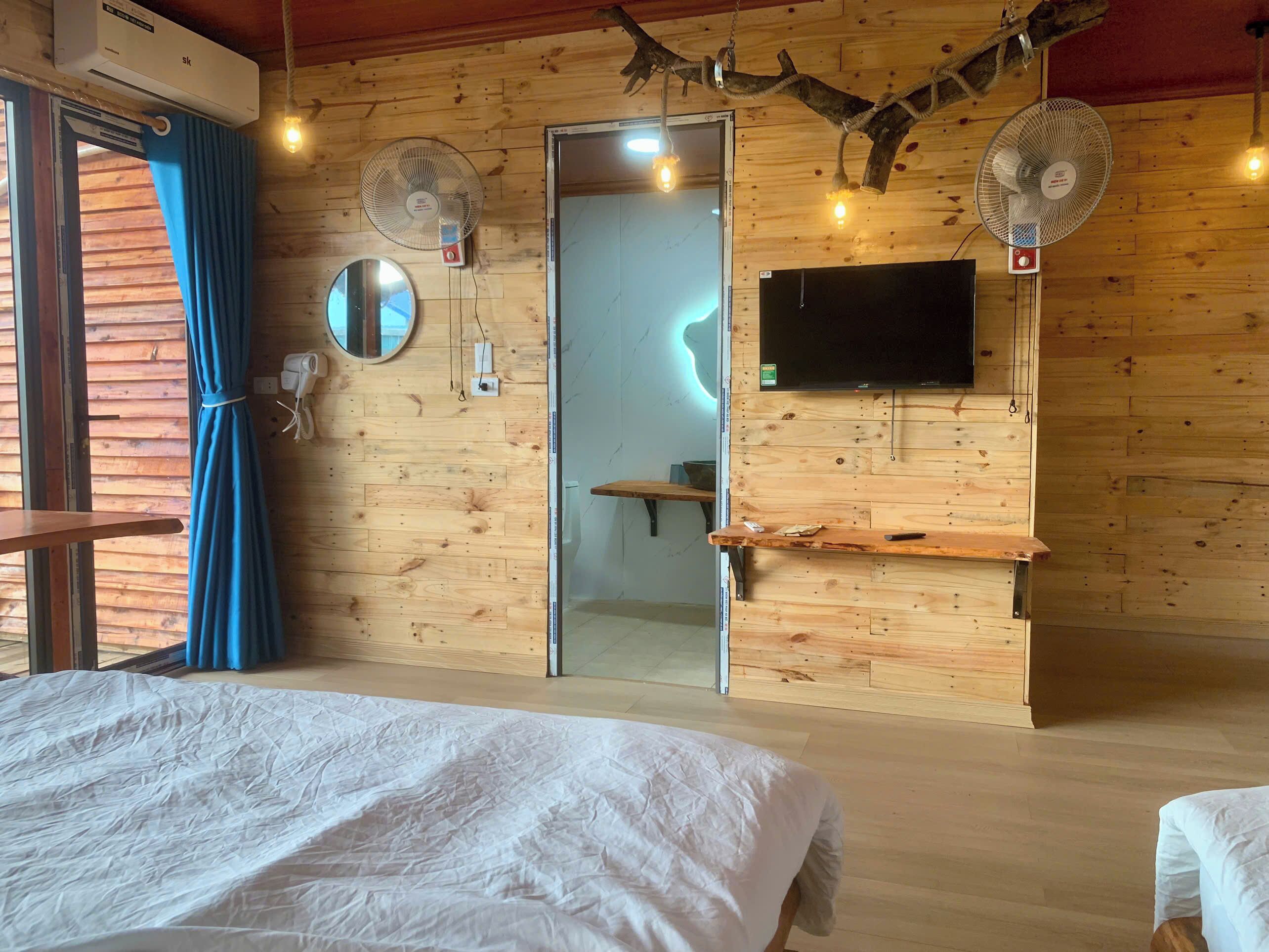 Ein Zimmer mit Holzboden, einem Bett, zwei Wandventilatoren, einem Fernseher und einer Tür zu einem weiteren Raum.