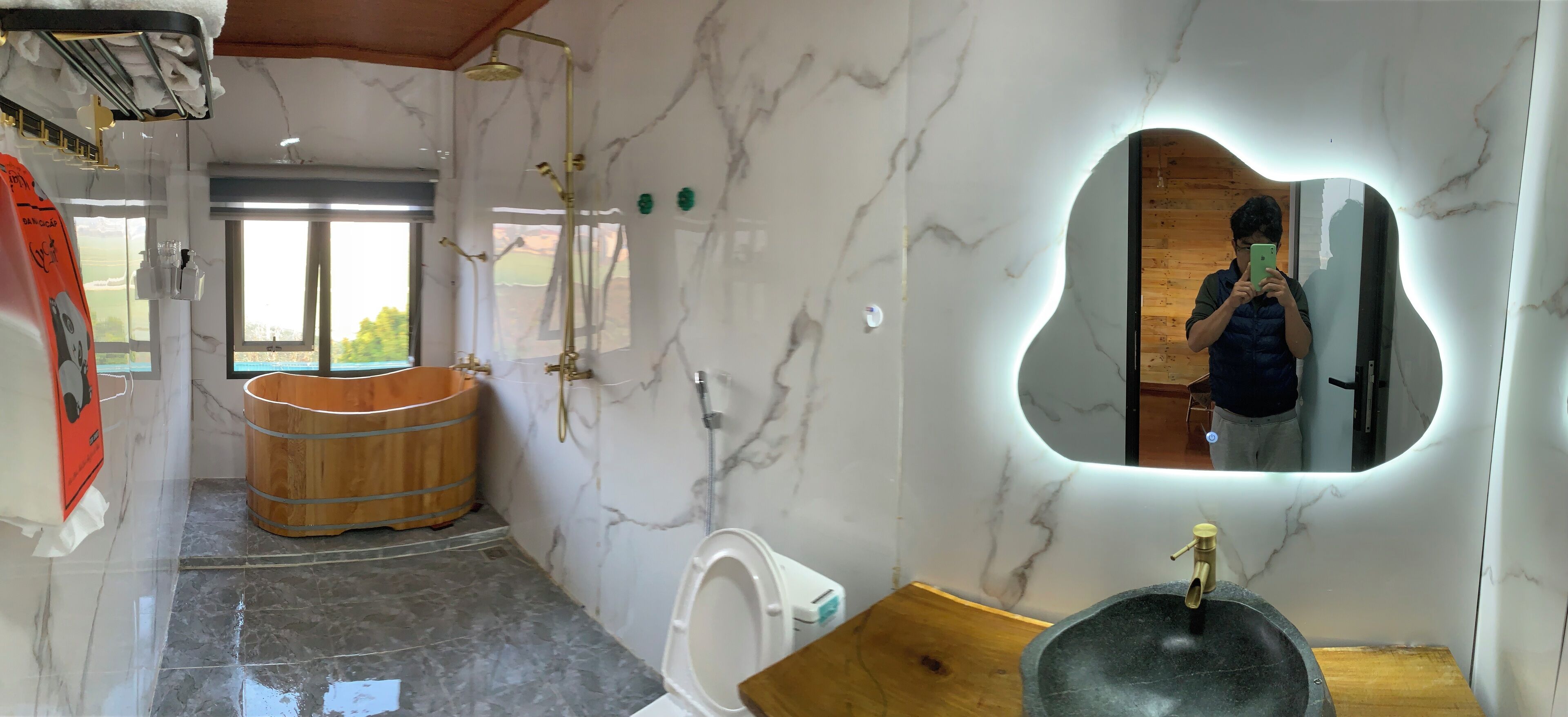 Un baño con pared de mármol, bañera de madera, espejo grande y lavamanos de piedra.