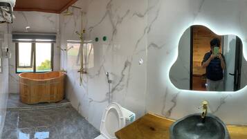 Un baño con pared de mármol, bañera de madera, espejo grande y lavamanos de piedra.