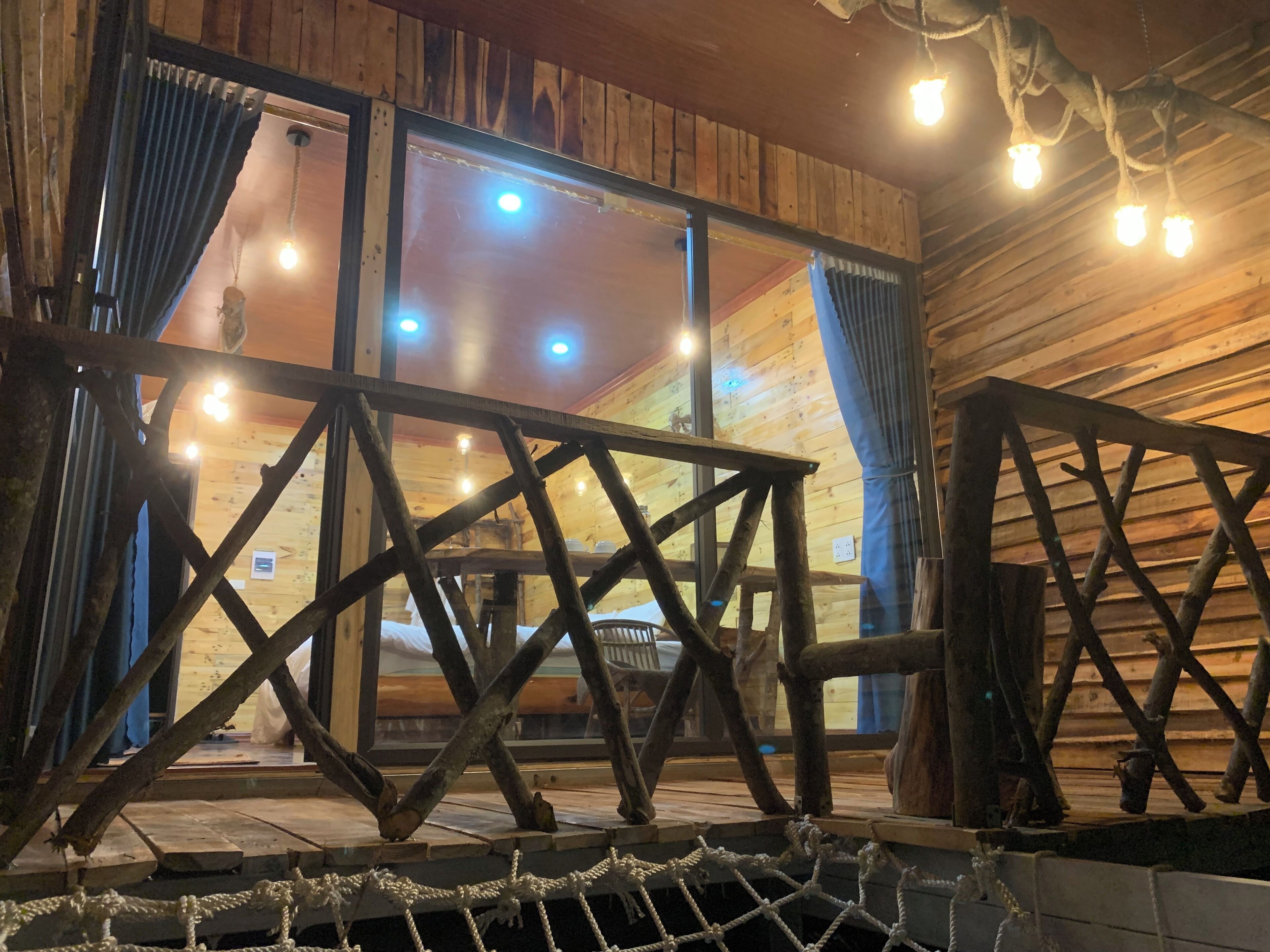 A rustic wooden cabin interior with a wooden deck, rope railing, and a large window with curtains.