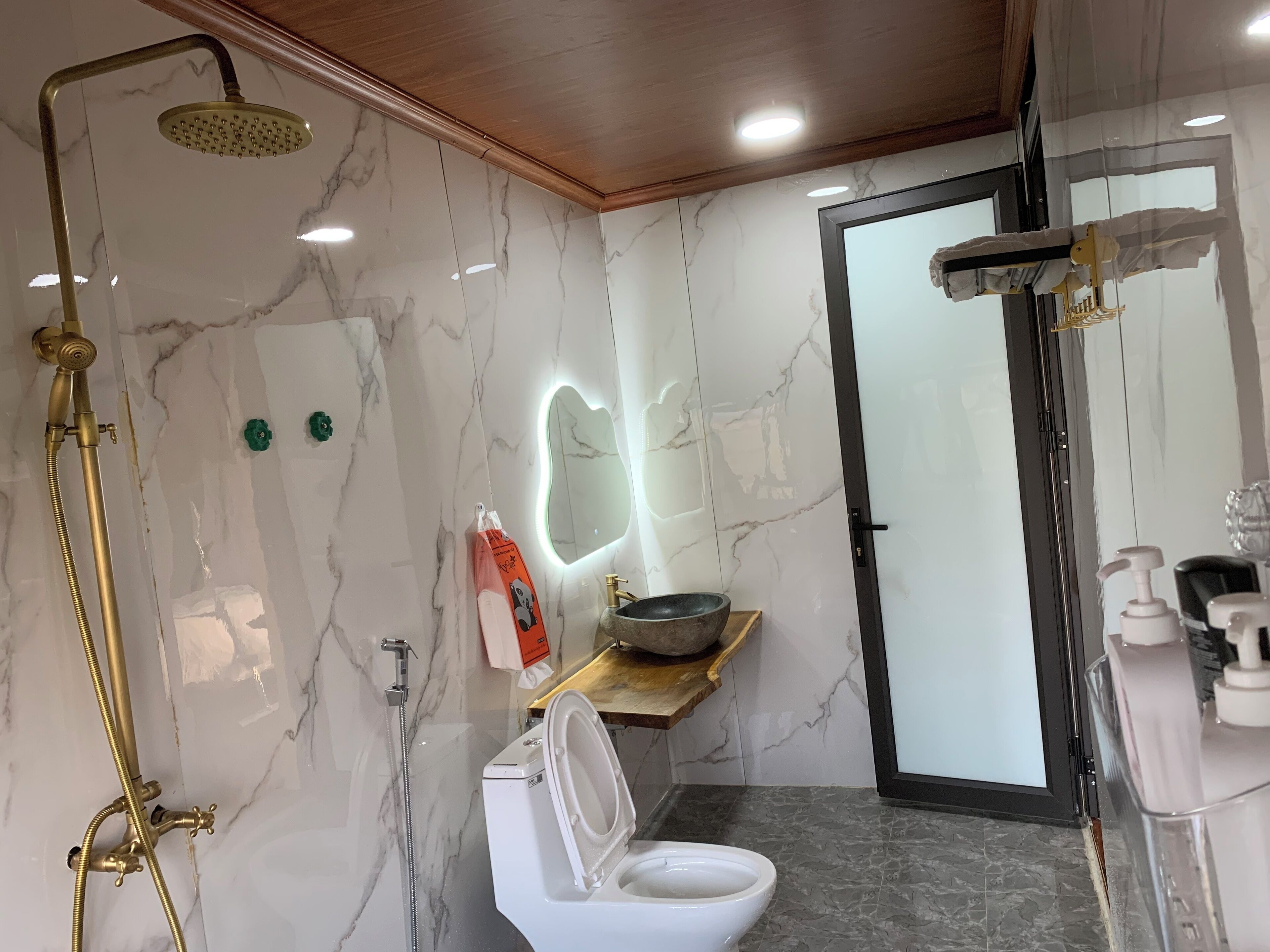Ein modernes Badezimmer mit Marmorwand, Toilette in Weiß, goldener Duschbrause und einem Waschtisch mit einer Schüssel.
