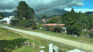 Une route de campagne bordée de maisons et d’arbres, un arc-en-ciel et des montagnes en arrière-plan.