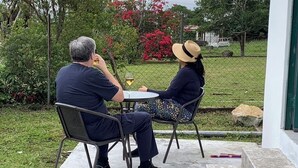 Deux personnes sont assises à une table à l’extérieur, devant une boisson, entourées de verdure et d’une clôture.