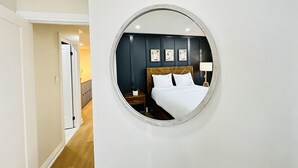 Una habitación de hotel con un espejo redondo en la pared, una cama con ropa blanca, un cabecero de madera, una mesita de noche con lámpara y una pequeña planta en maceta.