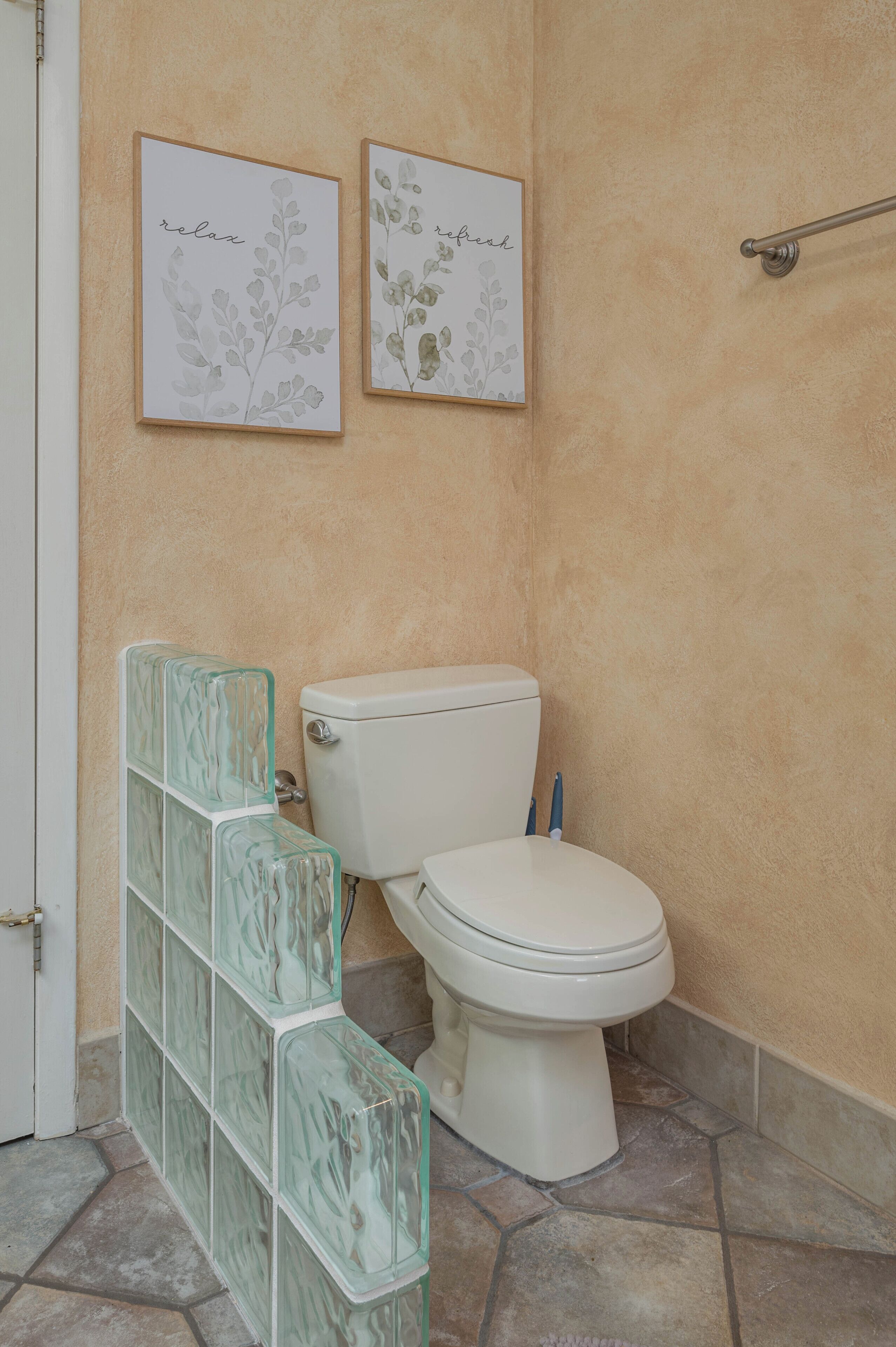 Un baño con inodoro, lavamanos de vidrio bloqueado y dos cuadros con motivos florales.