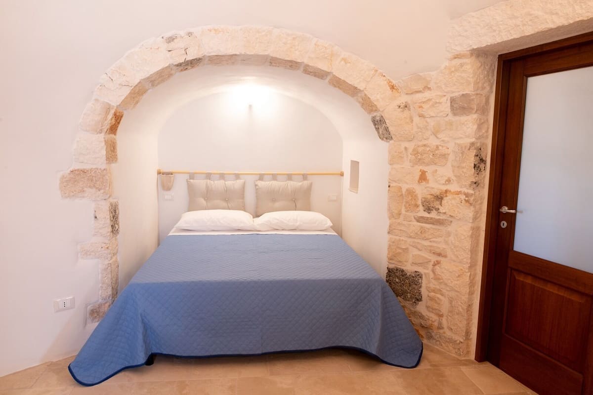 A bedroom with a stone archway, a bed with a blue bedspread, and a wooden door.