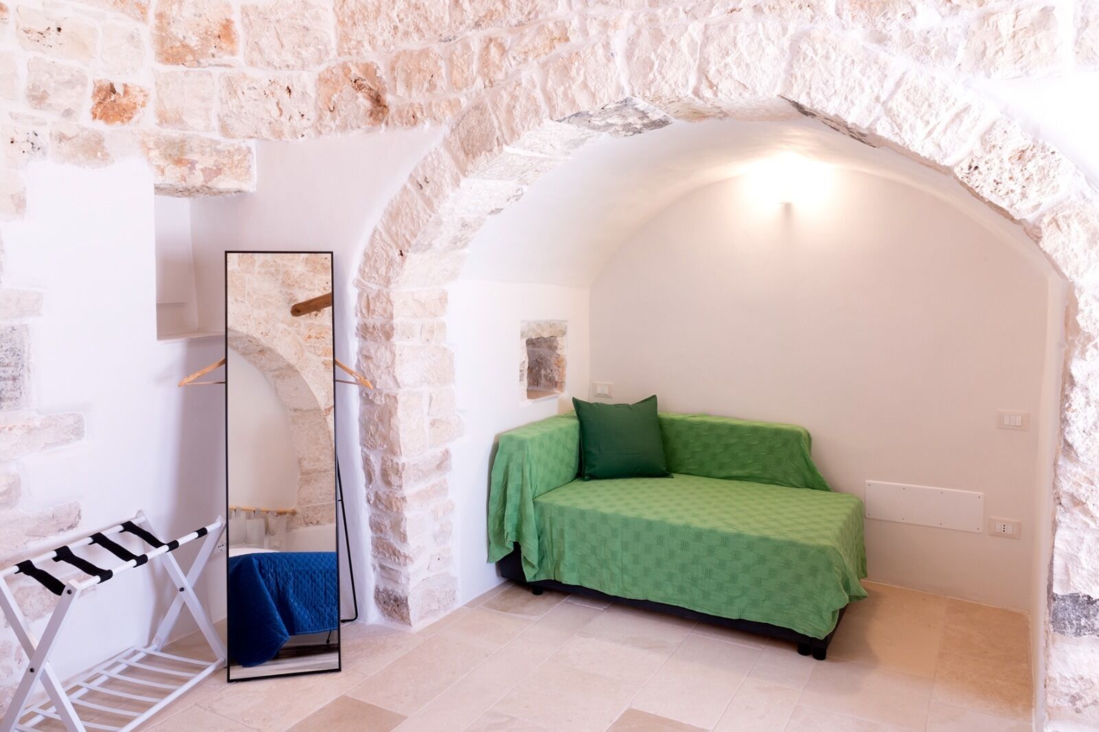 A room with a stone wall, a bed with green bedding, a mirror, and a drying rack with clothes.