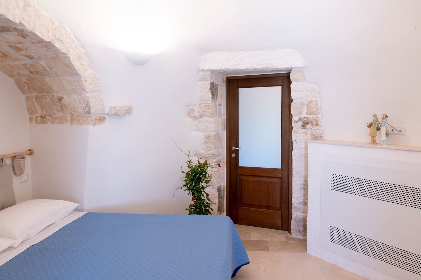 A bedroom with a bed, a wooden door, stone walls, a radiator, and a small plant.