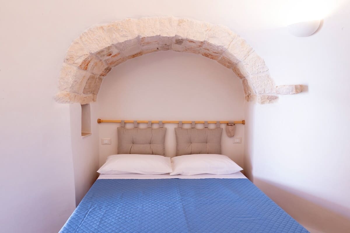 A single bed with a blue quilt, a wooden headboard, and a stone arch above it.