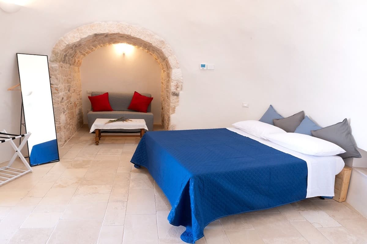 A bedroom with a bed, a sofa, a table, and a stone archway.