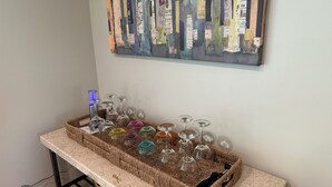 Zona de bar con una bandeja tejida que contiene copas y botellas de vino, una guitarra y una obra de arte en la pared.