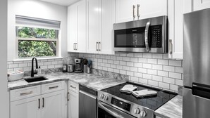 A modern kitchen with white cabinets, a stainless steel refrigerator, a microwave, an oven, and a window with a view of greenery.