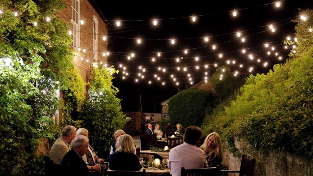 An outdoor dining area with string lights, surrounded by greenery and brick walls. - Chelsea Parkfields  (Ross-on-Wye)