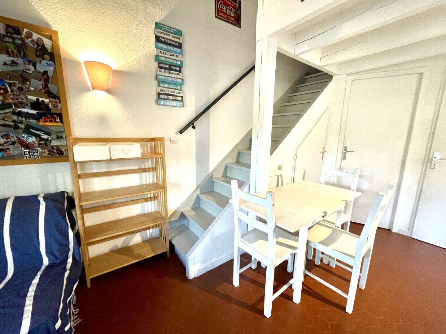 A room with a white staircase, a dining table with chairs, a wooden shelf unit, and a wall with a photo collage.