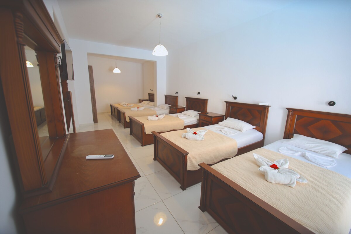 A hotel room with four single beds, each with neatly made linens and a towel with a flower. There is a wooden dresser with a remote control on it, and a large mirror on the left wall.