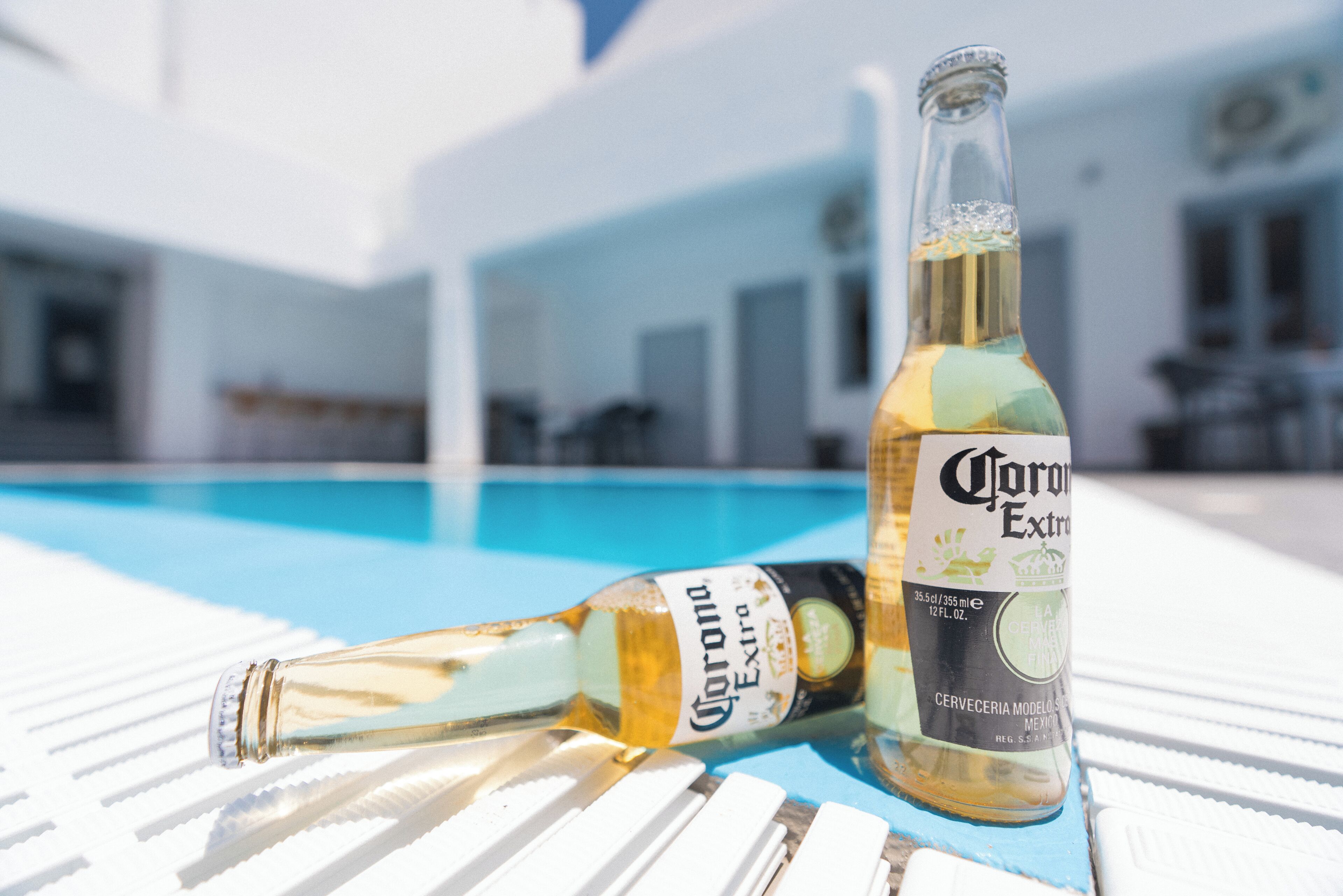 Twee flessen Corona Extra bier op een witte tafel bij een zwembad.