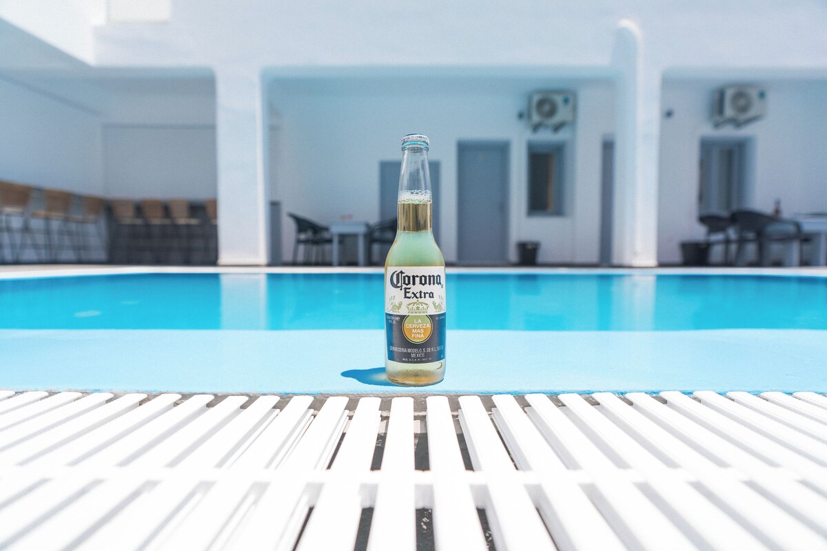 Une bouteille de bière Corona Extra posée sur une chaise blanche au bord de la piscine, avec une piscine et d’autres chaises longues en arrière-plan.