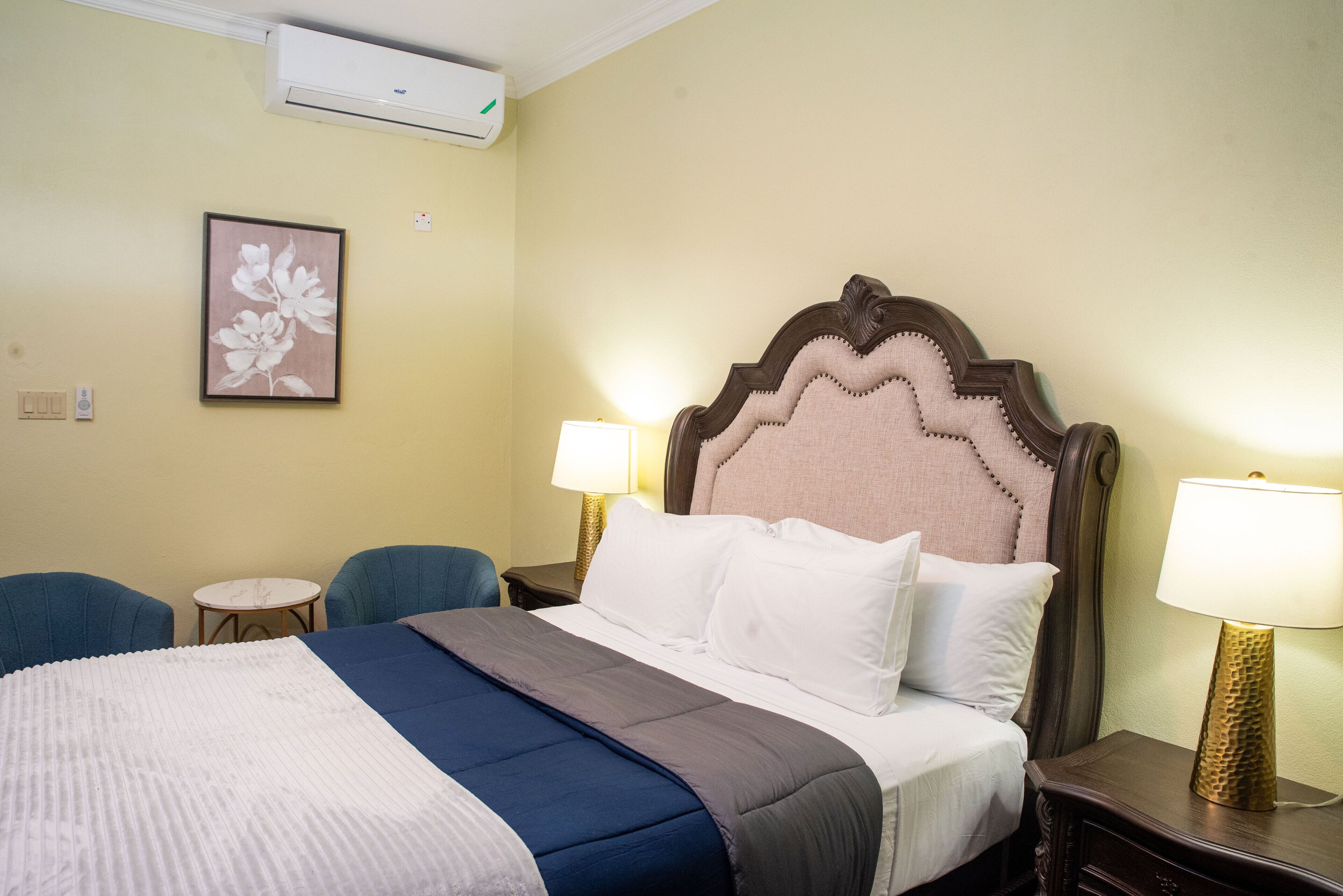 Une chambre d’hôtel comprenant un lit, deux chaises, une petite table, une lampe et un climatiseur fixé au mur.