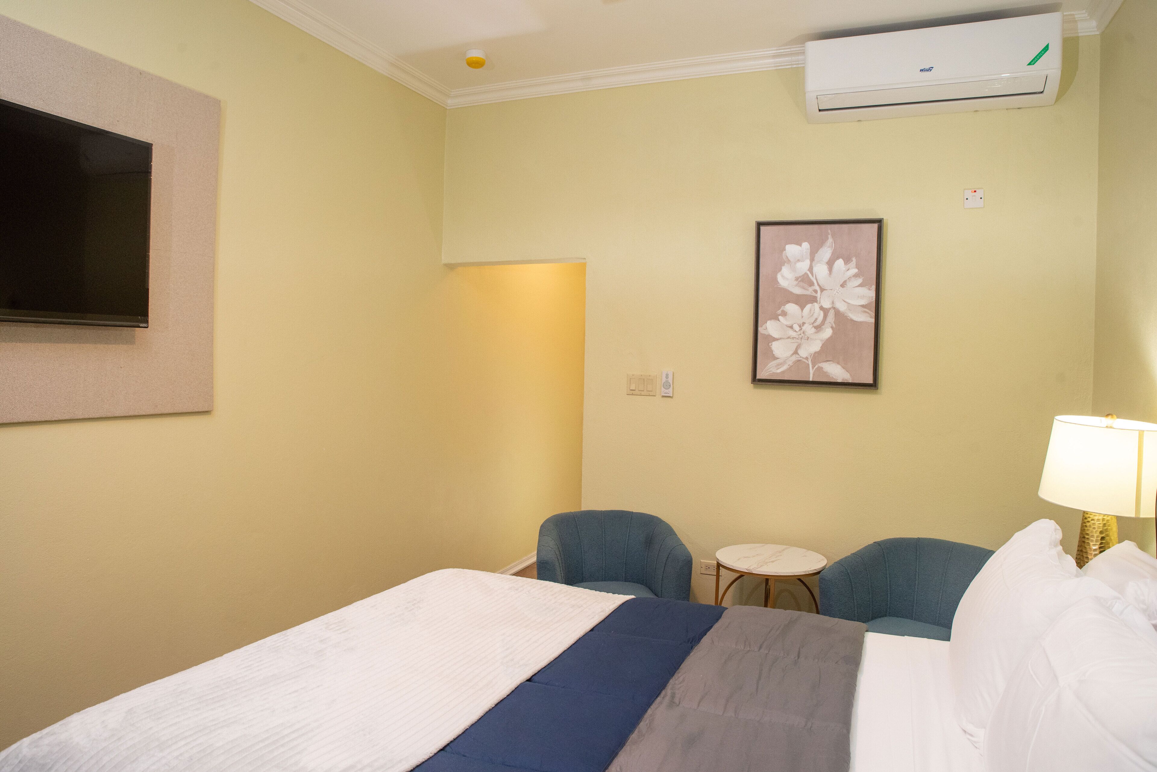 Een hotelkamer met een bed, twee stoelen, een tafeltje, een televisie, een airconditioner en een lijst aan de muur.