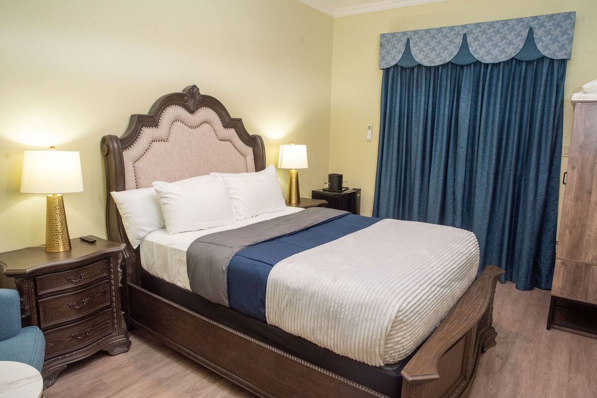 A neatly made bed with a headboard, two bedside tables with lamps, a blue curtain, and a wooden wardrobe.