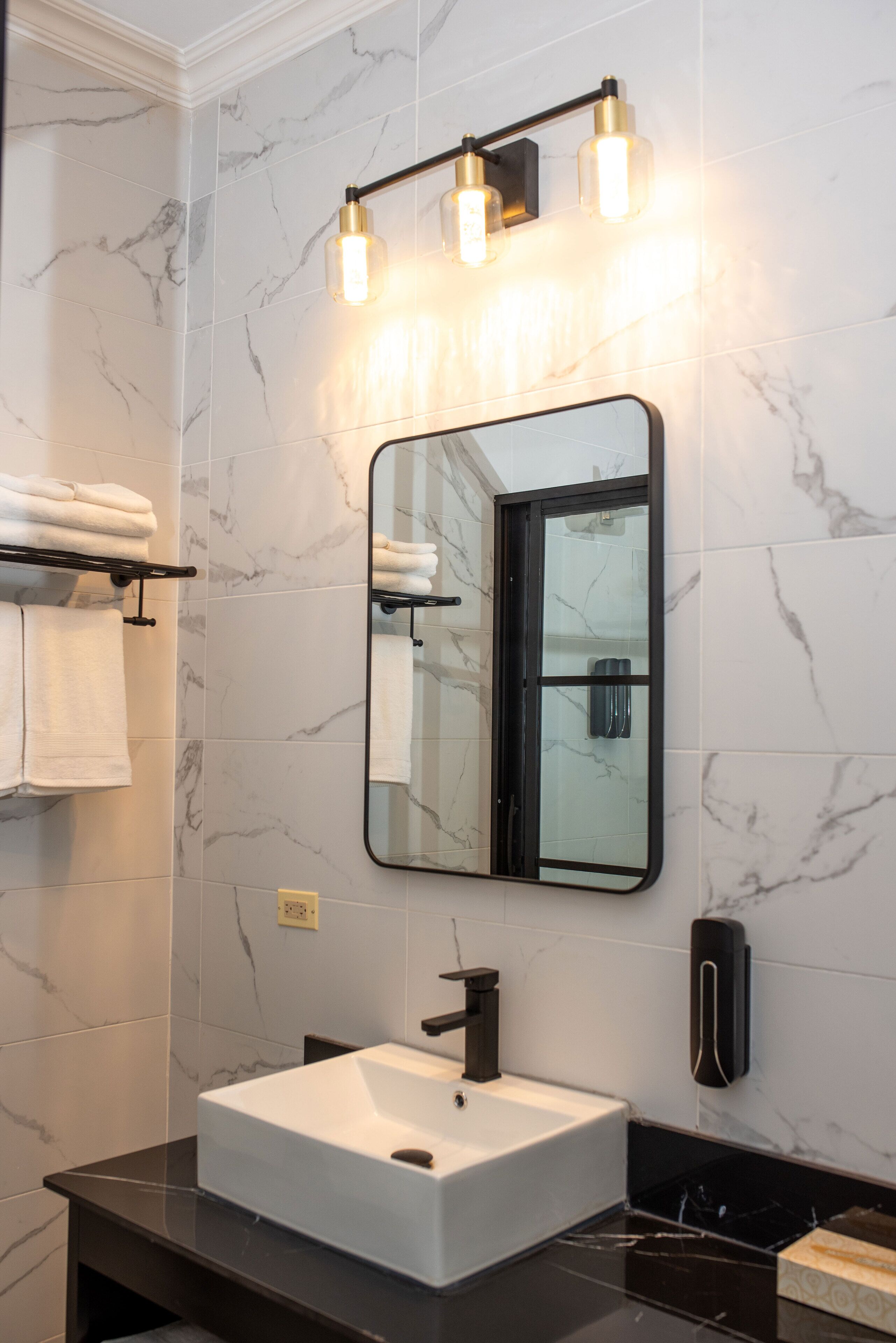 Un bagno con un lavandino bianco, un piano di lavoro nero e uno specchio con cornice nera. Asciugamani su uno stendino e un dispenser di sapone a parete.