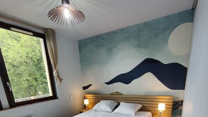 A bedroom with a wooden bed, a nightstand with a lamp, a window with curtains, and a wall mural.
