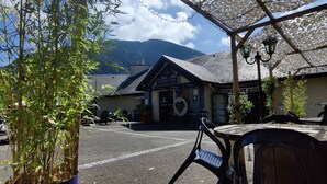 A covered outdoor seating area with a view of a mountain and a building with a sign reading "Café du Soleil."