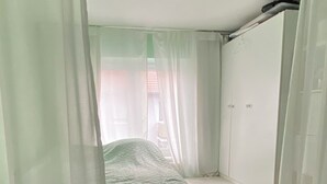 Um quarto pequeno, com cama de solteiro e dossel, paredes brancas e chão de madeira.
