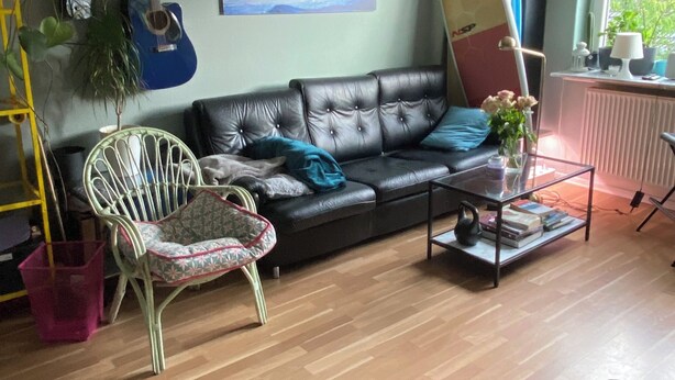 Uma sala de estar com um sofá de couro preto, uma poltrona de vime branco, uma mesa de centro de vidro e um prancha de surf na parede.