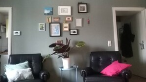 Sala de estar com duas poltronas de couro preto, uma com almofada branca e outra com almofada rosa. Há uma pequena mesa com uma planta em vaso no centro. A parede é decorada com várias imagens emolduradas e um termostato.