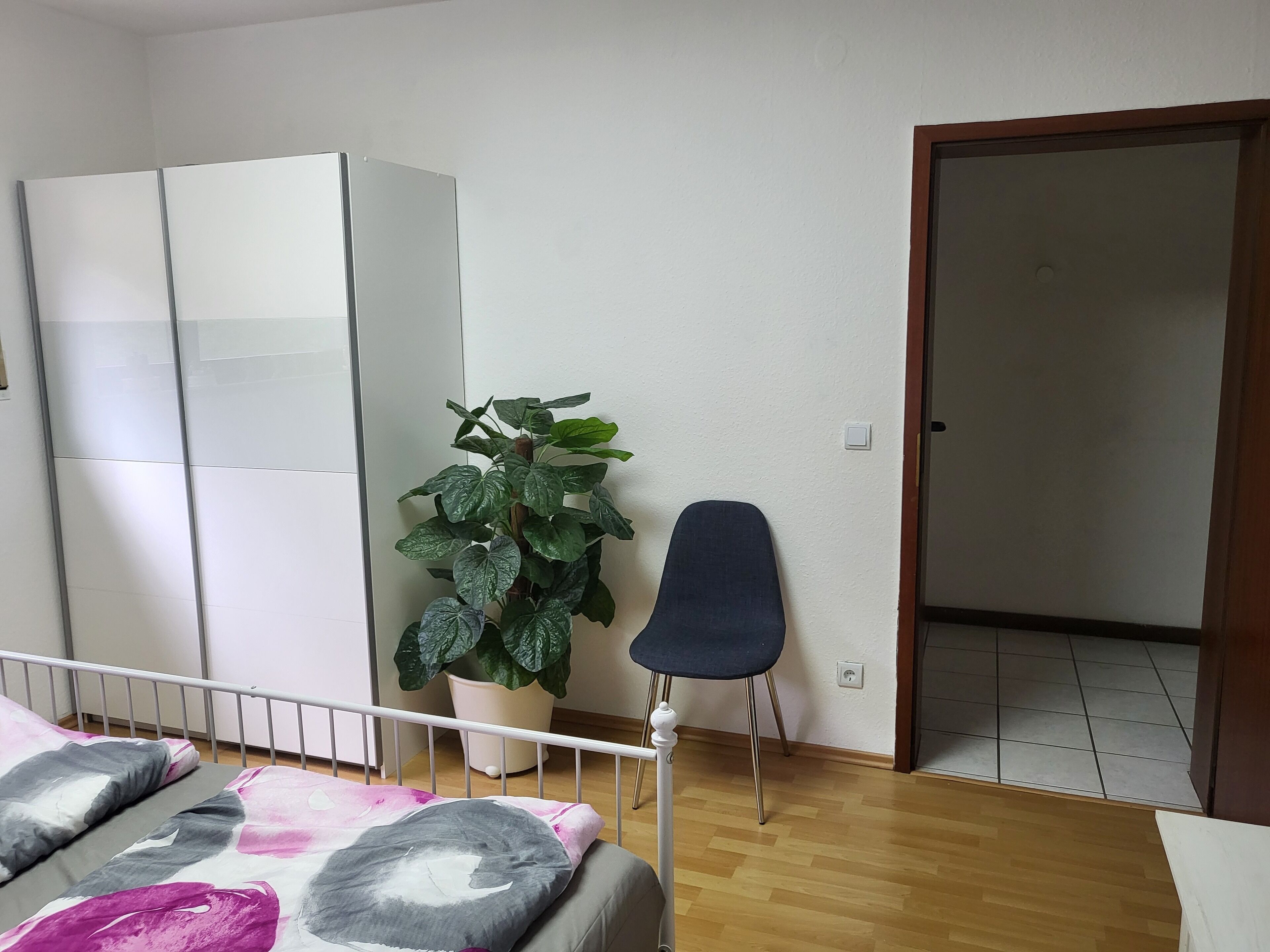 Une chambre à coucher comprenant un lit, une armoire, une plante en pot, une chaise et une porte donnant sur une autre pièce.