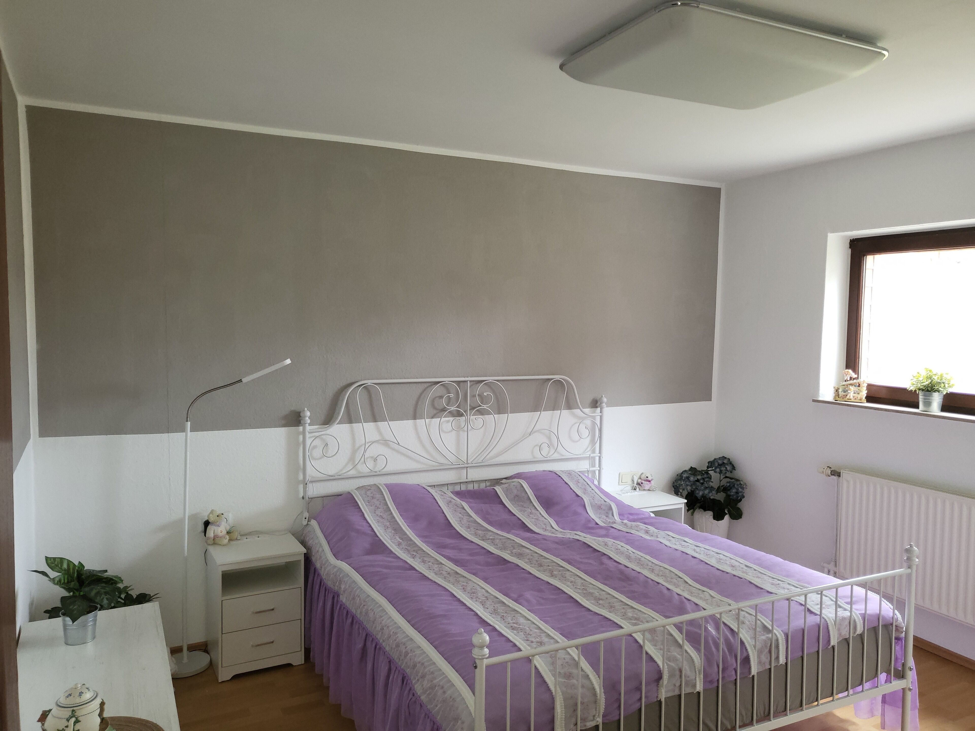 Ein Schlafzimmer mit Bett, Nachttischen, Fenster mit Pflanze und Heizkörper.