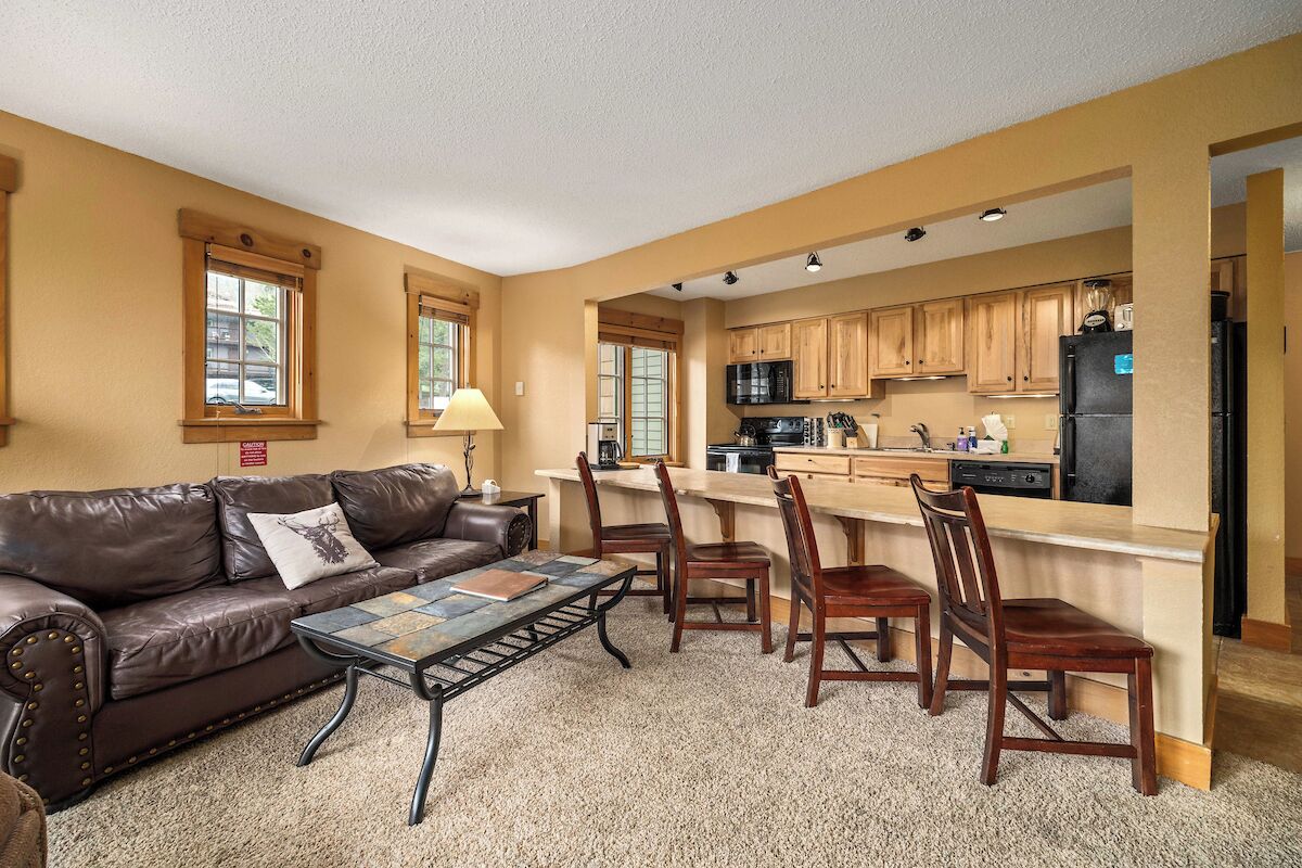 A spacious living room with a brown leather couch, a coffee table, and a dining area with wooden chairs and a kitchen in the background.