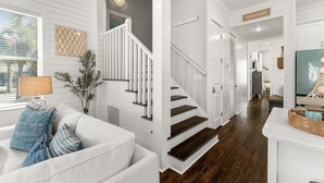 A modern interior with a white staircase, a living area with a white sofa and blue cushions, and a wooden floor. - Sailfish Lookout, Private Beach Access (Panama City Beach)