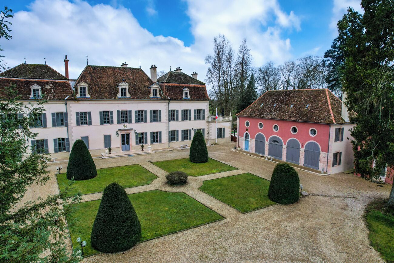 Un grand bâtiment historique à l’architecture symétrique, doté d’une cour centrale avec des espaces verts aux formes géométriques et des haies taillées.