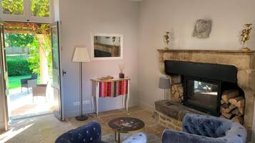 Een woonkamer met een open haard, twee gestoffeerde fauteuils, een bijzettafeltje, een lamp en uitzicht op de tuin door een open deur.