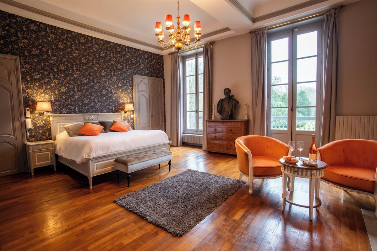 Een slaapkamer met een groot bed, twee nachtkastjes, een dressoir, een kroonluchter en een raam met gordijnen.